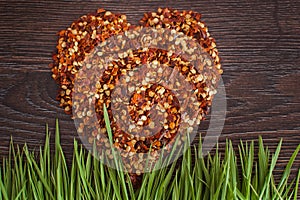 Red hot chili pepper flakes lying on wooden table in heart shape with green grass. Cooking concept, seasonings for food.