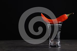 Red hot chili pepper and vodka in shot glass on grey table against black background, space for text