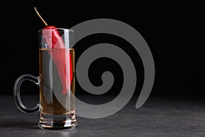 Red hot chili peppers and vodka in shot glass on grey table against black background, space for text