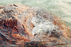 Red huge large stone rock in water on beach. Closeup of sea ocean lake wave splash with white foam.