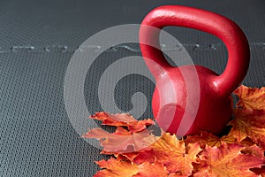 Red iron kettlebell on a black rubber gym floor, with orange and yellow maple leaves, fall fitness