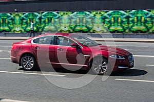 Red Jaguar XF riding on the highway. Side view of shiny compact executive car driving in the street