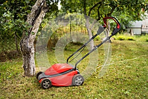 Red lawn mower outdoor in the backyard. green grass and fruit trees background. Gardening concept.