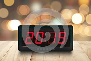 Red led light illuminated numbers 2019 on digital alarm clock face on wooden table with defocused colorful Christmas lights bokeh