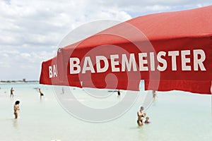 Red lifeguard umbrella on a beach against blue sky. Some bathers ont he background. The text on the umbrella Bademeister