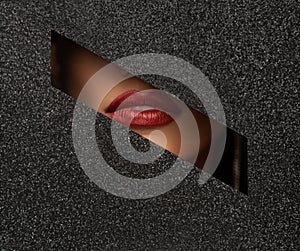 Red lips peep through a hole in shiny black paper.