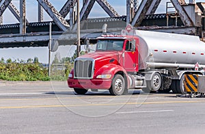 Red local big rig semi truck with day cab transports fuel in the tank trailer running under the old bridge