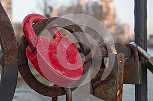 Red, massive metal padlock hanging from round, rusted element of bridge across river, among other locks that were rusted