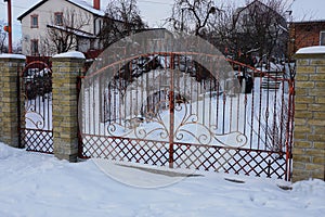 Red metal gates of forged rods with a pattern and part of a brick fence on the street in white snow