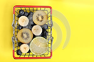 Red metal shopping basket full of fruits and berries isolated on a yellow background. Lime, banana, blueberries, kiwi and apples