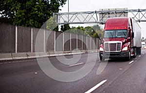 Red modern big rig semi truck with trailer running on wide highway in raining weather