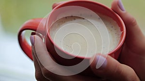 Red mug of cappuccino in female hands on the background of the window