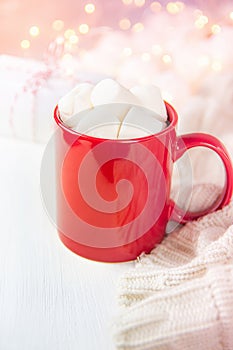 Red Mug with Hot Chocolate Cocoa Drink and Marshmallows on Top. Glittering Sparkling Garland Lights in Background. Knitted Sweater
