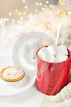 Red Mug with Hot Chocolate Cocoa Drink and Marshmallows on Top Mince Pie. Sparkling Garland Lights in Background. Knitted Sweater