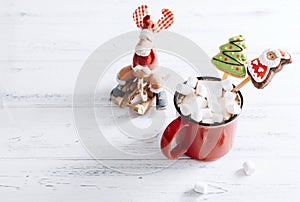 Red mug of hot chocolate with marshmallows and cookies, toy reindeer on a sleigh ,red Christmas balls on a white  wooden