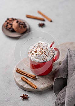 Red mug of hot cocoa or chocolate with whipped cream and cookies, cinnamon sticks on a gray background