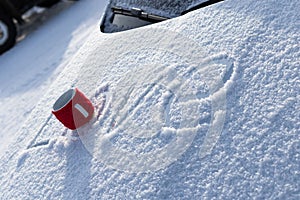 A red mug with a hot drink on a snowy car hood on a bright winter sunny day. The inscription on the snow is love. Selective focus