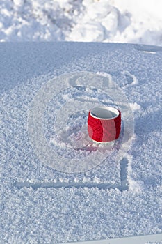 A red mug with a hot drink on a snowy car hood on a bright winter sunny day. The inscription on the snow is love. Selective focus.