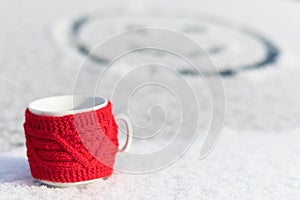 A red mug with a hot drink on a snowy car trunk on a frosty sunny winter day. On the rear window of the car, a drawing of a smilin