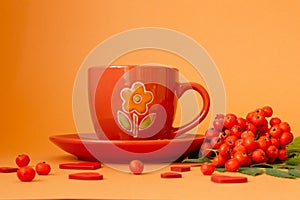 Red mug with a saucer and ripe rowan berries on an orange background. Cozy autumn concept
