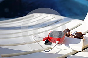 Red music portable speaker is charged from the power bank via usb on a deck chair near the pool. Concept is always in touch,