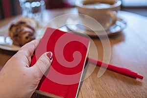 A red note and red pen is on the table in a cafe, a cup of coffee and a croissant on background. Female hand holding a