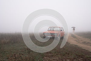 Red old retro car near the dirt road, young girl in white coat under umbrella walks in the fog on the background