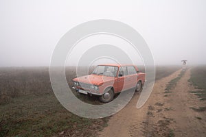 Red old retro car near the dirt road, young girl in white coat under umbrella walks in the fog on the background