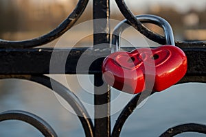 Red padlock is composition of two hearts hanging on black ornate railing of bridge fence on blurred river background