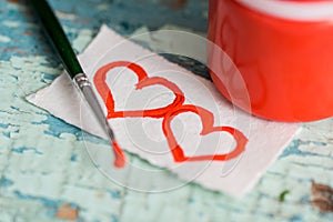 Red paint gouache in a jar, brush. painted two hearts on a scrap of paper. on a textured old cracked blue green background on a wo