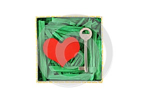 Red painted wooden heart and metal key enclosed with green paper raffia strips in paper box isolated on white background