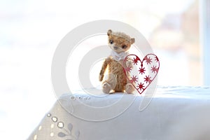 Red paper heart and the small brown toy bear made of wool sitting on a white napkin. warm colors. soft focus.