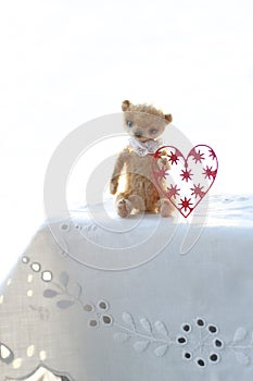 Red paper heart and the small brown toy bear made of wool sitting on a white napkin warm colors. soft focus.