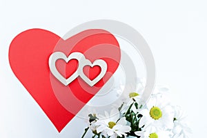 Red paper heart, two carved wooden hearts on it and white chrysanthemums on white background.