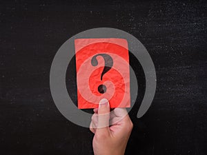 A red paper note with a question mark being held in a hand against a blackboard