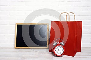 Red paper shopping bags with blank blackboard and alarm clock on white brick wall background