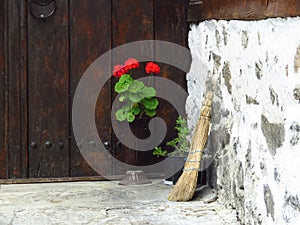 Red pelargonium geranium flowers and a broom against brown wooden door and stone wall. Traditional folklore concept.