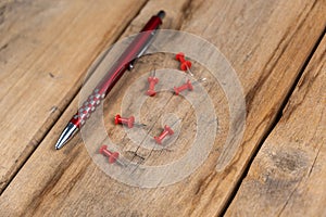 A red pen and a push pin are against the wooden background. A metal ballpoint pen and six push pins lie on top of old cracked