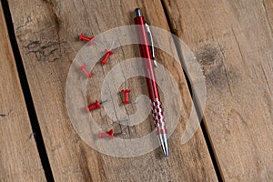 A red pen and a push pin are against the wooden background. A metal ballpoint pen and six push pins lie on top of old cracked