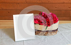 Red peony flowers in wicker basket and white frame on wooden table. Selective focus. View with copy space