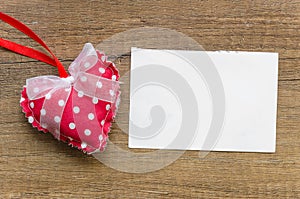 Red pillow heart with white dots and a bow and on wooden background with a blank list for greeting card. Space for text