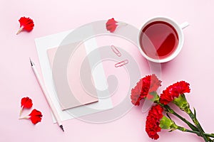 Red-pink objects. Cup of tea, carnation flowers Notepad for text on pastel pink background. Copy space. Top view Flat lay