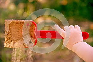 Red plastic scoop scoop in a children`s hand. Play in the sandbox. With a scoop of sand falls. Symbol of childhood and fun