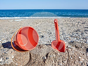 Red plastic shovel and bucket for children in the sand on the beach in front of the sea