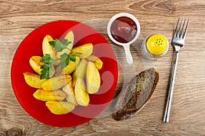 Red plate with slices of fried potatoes, parsley, sauce boat with ketchup, pepper shaker, fork, piece of bread on table. Top view