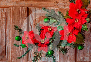 Red Poinsettias and bells decorate a Christmas tree branch against weathered door