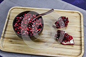 Red pomegranate seeds close up in a wooden bowl with a wooden spoon. Two pieces of pomegranate with ripe red seeds on a wooden