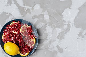 Red pomegranate seeds on yellow lemon slices in a blue ceramic plate on a textured gray concrete background