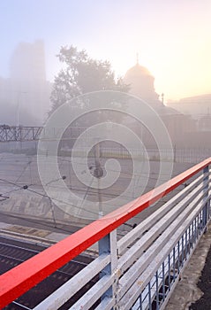 Red railings of a pedestrian bridge on the background of a temple in the fog in Novosibirsk