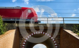 Red railway train is driving fast over a road bridge with an arch and red-white markings, intended motion blur
